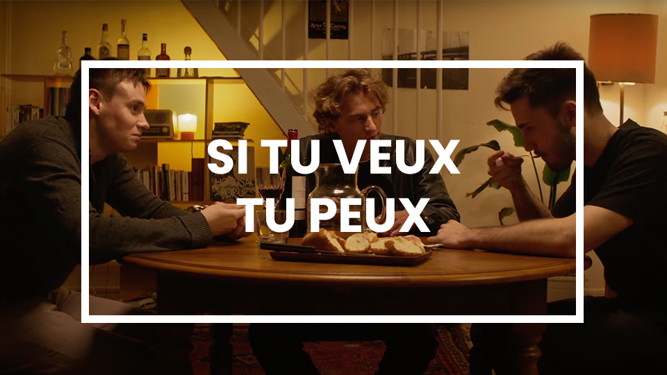 Trois jeunes hommes sont assis autour d’une table en bois dans un salon à l’éclairage tamisé et chaleureux. L’un d’eux mange, tandis que les deux autres semblent engagés dans une conversation sérieuse. Sur la table, une théière, des verres de vin et une assiette de pain apportent une atmosphère conviviale. En arrière-plan, une bibliothèque remplie de livres et de bouteilles, ainsi qu’un escalier menant à un étage supérieur. Le texte “SI TU VEUX TU PEUX”, écrit en blanc et encadré, renforce le message motivant de la scène.
