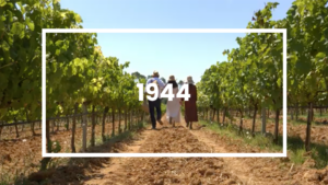 Quatre personnes marchant dans un vignoble sous un ciel bleu, entourées de vignes luxuriantes. Le chiffre '1944' est superposé au centre de l’image, encadré par un rectangle blanc.