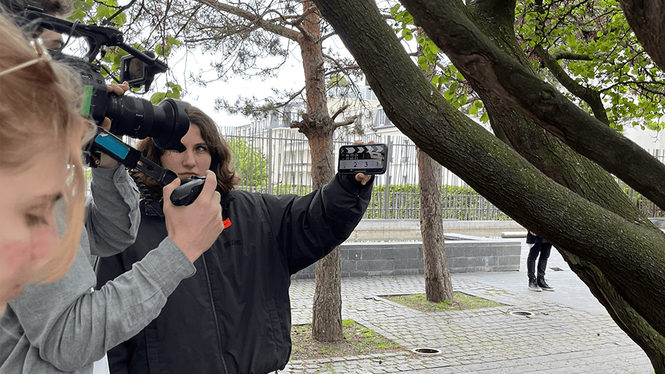 Sur un tournage en extérieur, une jeune femme habillée en noir tient un clap numérique, marquant le début d’une prise. À ses côtés, une équipe de tournage manie une caméra professionnelle et un micro perche, capturant chaque détail de la scène. Les arbres encadrent le décor urbain, ajoutant une touche de nature à cette ambiance de plateau de cinéma en plein air.