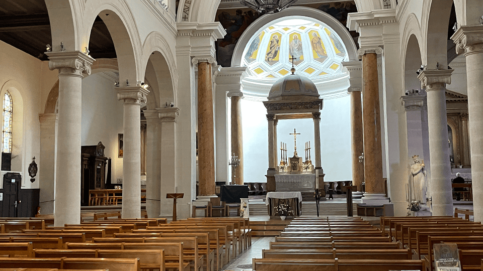 Intérieur d'une église majestueuse avec des arches élégantes, des colonnes en pierre et un autel central illuminé par la lumière naturelle. L’espace sacré est baigné d’une atmosphère paisible, avec des bancs en bois soigneusement alignés face à l’autel orné de chandeliers et d’une croix.