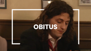 Un homme aux cheveux longs et bouclés, vêtu d’un costume sombre et d’une cravate rouge desserrée, est assis dans un bar ou un café. Il regarde vers le bas avec une expression pensive et mélancolique. L’ambiance est feutrée, avec des cadres accrochés aux murs en arrière-plan. Le texte 'OBITUS' est superposé au centre, encadré par un rectangle blanc.