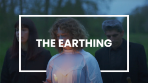 Trois personnes marchant dans un cadre naturel au crépuscule. Une femme en robe blanche tient une bougie allumée, entourée de deux autres individus vêtus de noir. Le texte 'THE EARTHING' est superposé au centre de l’image, encadré par un rectangle blanc.