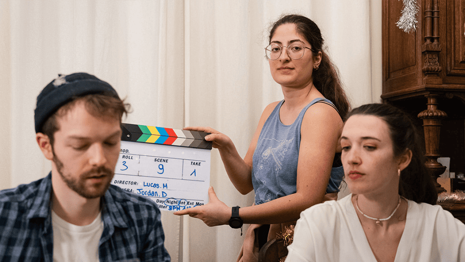 Membre de l’équipe technique tenant un clap de tournage avant une prise, entourée de deux comédiens concentrés sur leur rôle.