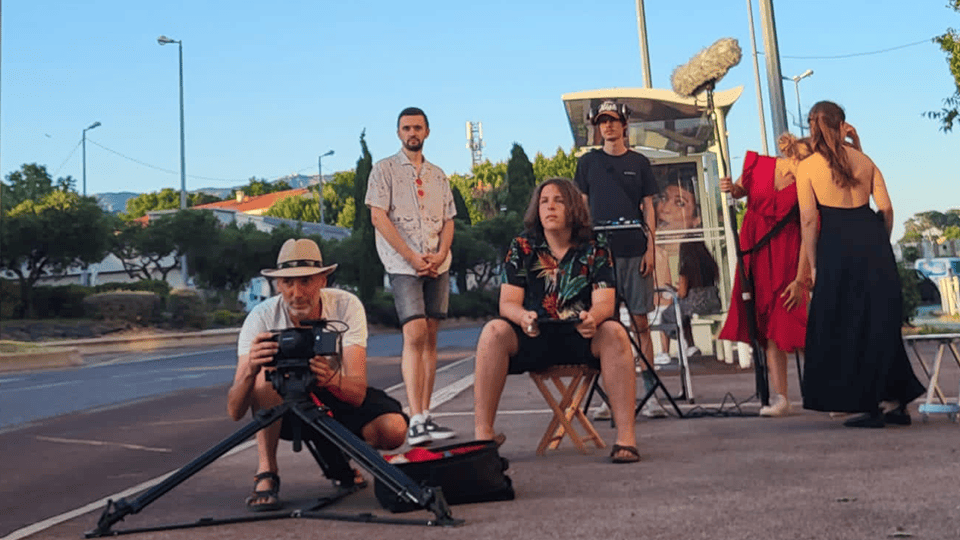 Une équipe de tournage en plein air est en action sur un trottoir bordant une route. À l’avant-plan, un caméraman portant un chapeau de paille est accroupi, manipulant une caméra montée sur un rail pour un travelling. Au centre, une personne aux cheveux longs, assise sur un tabouret, semble donner des directives ou se préparer à une scène. Derrière elle, un homme en chemise imprimée et short observe la scène. Plus loin, un perchiste tient un micro au-dessus d’un abri de bus, où un écran de contrôle est installé. Sur la droite, deux personnes sont en discussion, dont une en robe rouge avec une ceinture noire et l’autre en tenue sombre, de dos. L’environnement urbain est baigné d’une lumière chaude, évoquant une fin d’après-midi ou un début de soirée.