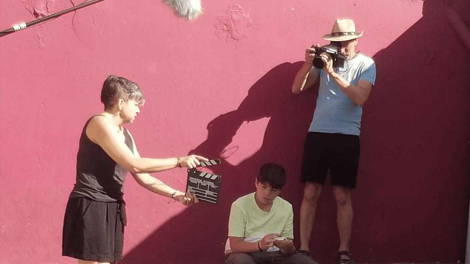 Tournage d’une scène de film en extérieur : une femme utilise un clap de cinéma devant un adolescent assis au sol, pendant qu’un cameraman en chapeau filme la scène contre un mur rose vif.