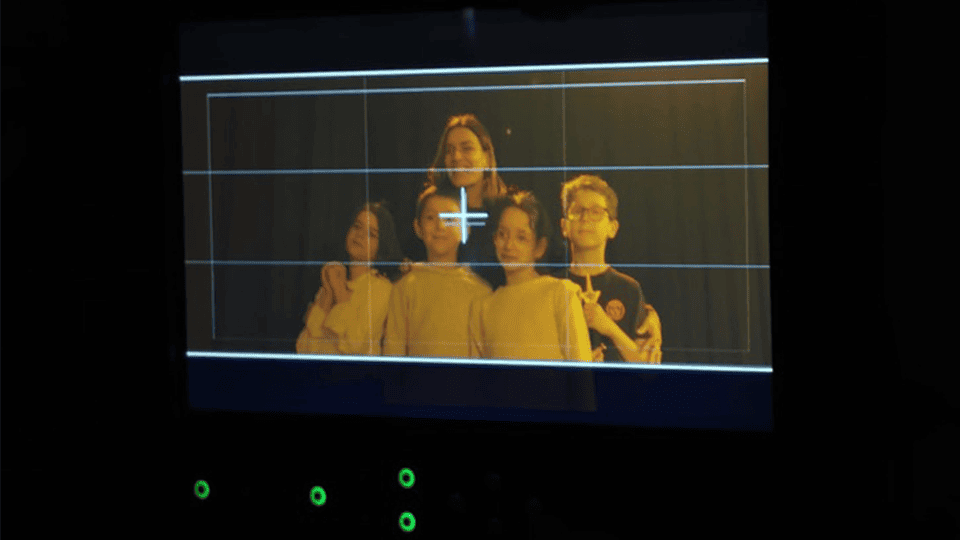 Vue de l’écran d’un moniteur de tournage affichant un groupe d’enfants et une adulte posant sous une lumière chaude, encadrés pour une prise de vue en studio.