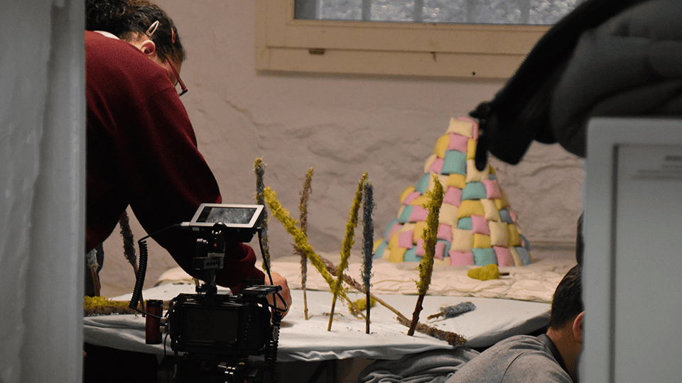 Mise en place d’une maquette miniature pour le tournage du clip “Please Babe” du groupe Polycool, avec une pyramide pastel en fond et une caméra de cinéma en premier plan.