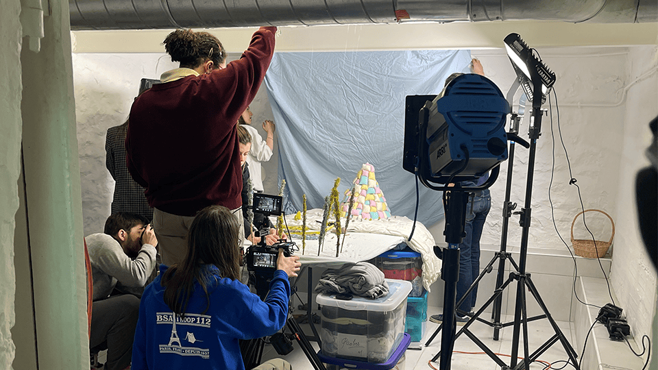 Coulisses d’un tournage en studio avec plusieurs membres de l’équipe technique réglant les caméras et l’éclairage, devant une maquette sur une table, dans un décor blanc.