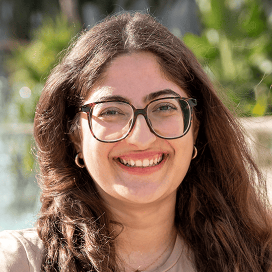 Portrait d’une jeune femme souriante portant des lunettes et un t-shirt beige, avec de longs cheveux bruns ondulés. L’arrière-plan montre un décor extérieur ensoleillé avec de la verdure et une fontaine floue.