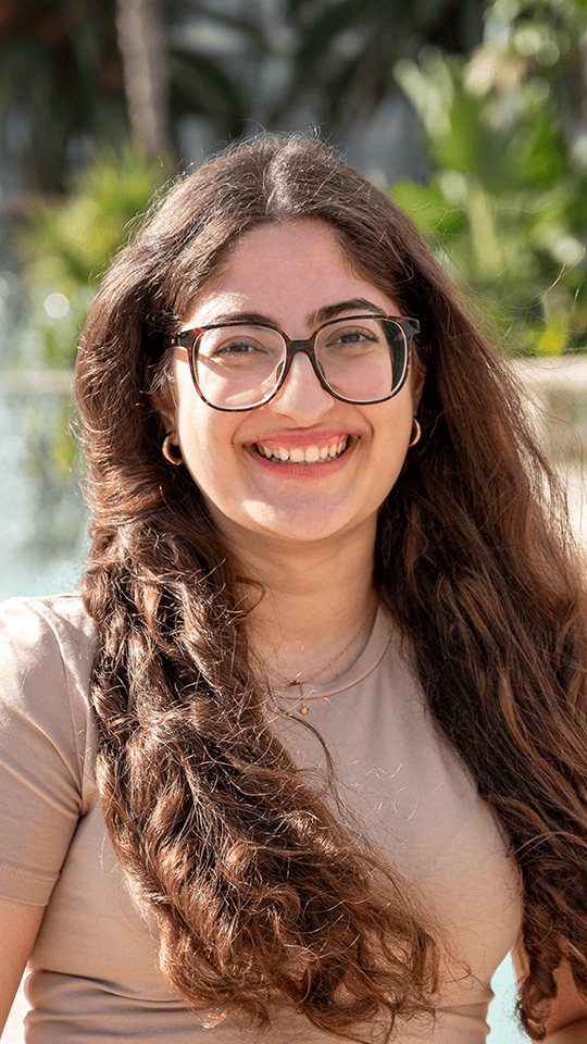 Portrait d’une jeune femme souriante portant des lunettes et un t-shirt beige, avec de longs cheveux bruns ondulés. L’arrière-plan montre un décor extérieur ensoleillé avec de la verdure et une fontaine floue.