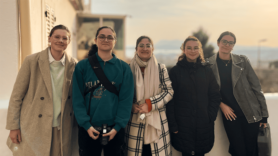 Cinq jeunes femmes posent côte à côte en extérieur, souriantes, sous une lumière dorée de fin de journée. Elles sont vêtues de manteaux d'hiver et regardent l'objectif, avec une vue floue de la ville et des montagnes à l'arrière-plan.